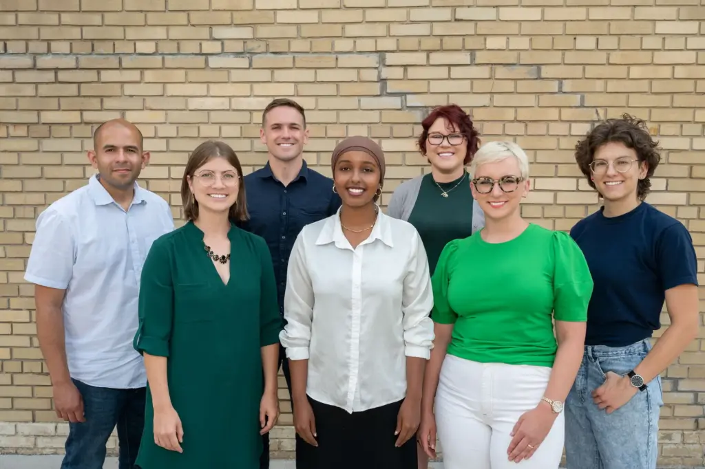 Posed group photo of 7 people smiling with a tan brick wall behind them.