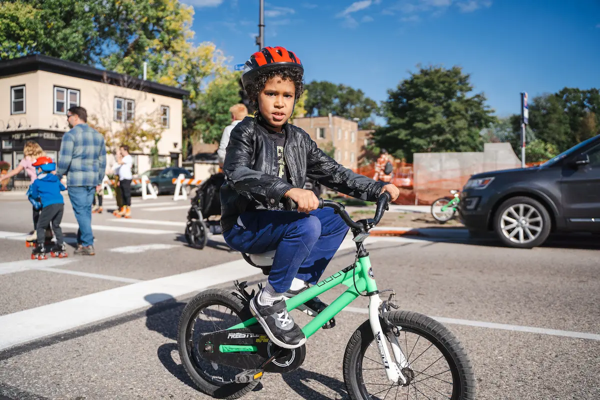 Young kid on bicycle with helmet, parent and other child on roller skates in the background.