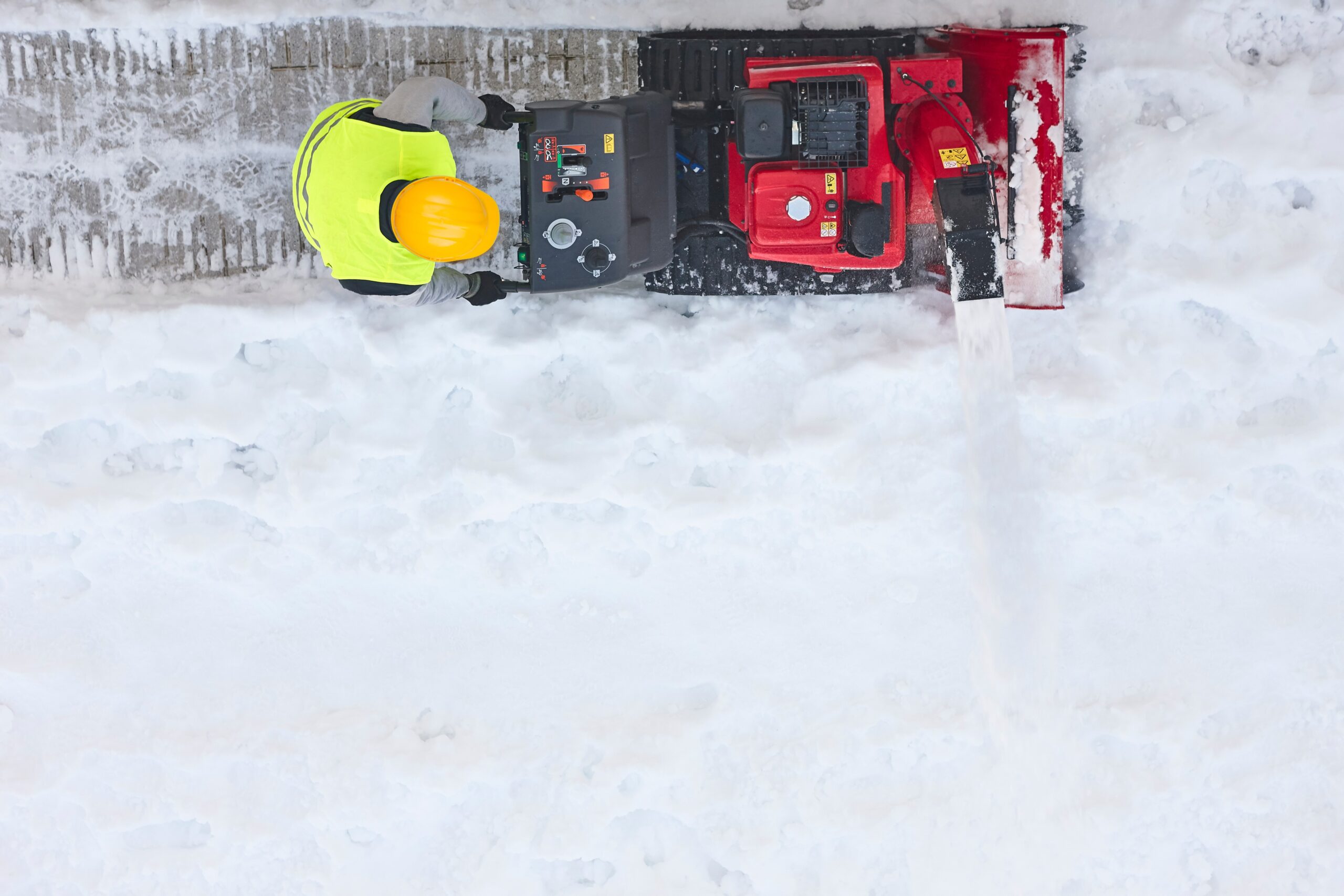 Aerial shot of a person using a motorized plowing machine to clear snow on the sidewalk.