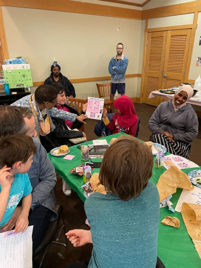 A diverse group of people gather around a table, looking at a drawing made and held up by a young girl with brown skin wearing hijab.