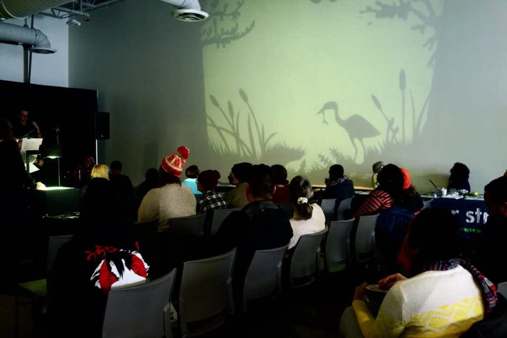 Dark theatre room with audience members heads turned to the screen. On the screen is a puppet show, "How Did We Get Here?".