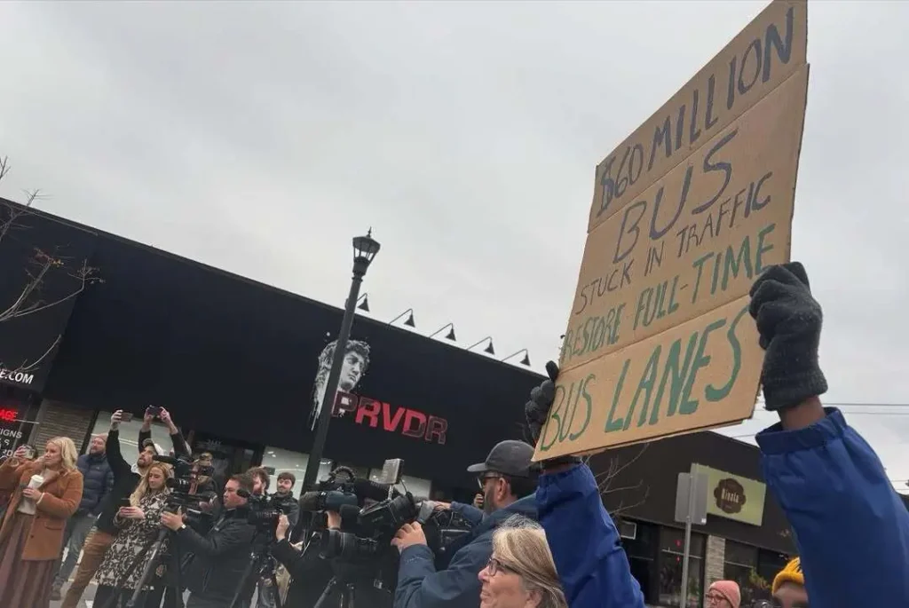 Someone holding a sign above a crowd reading "60 Million Bus Stuck in Traffic - Restore Full-Time Bus Lanes"
